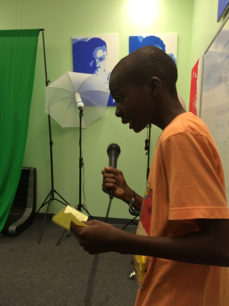 A youth looking at a sticky note and holding and speaking into a microphone in a green room