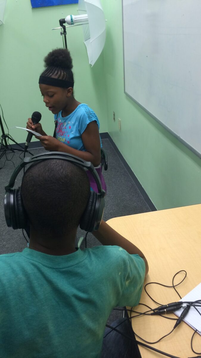 A youth wearing headphones sitting at a classroom desk listening to another youth holding and speaking into a microphone