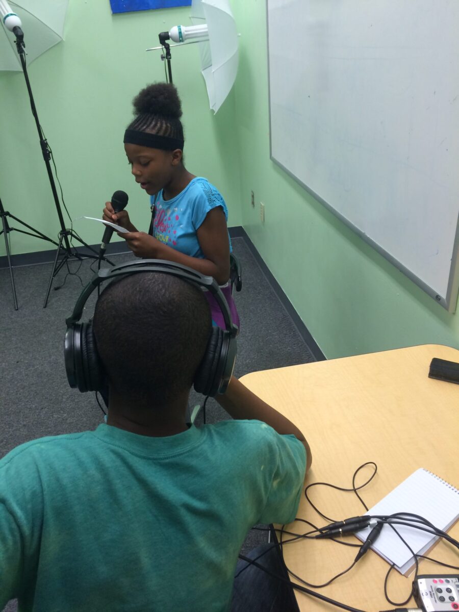 A youth wearing headphones sitting at a classroom desk listening to another youth holding and speaking into a microphone