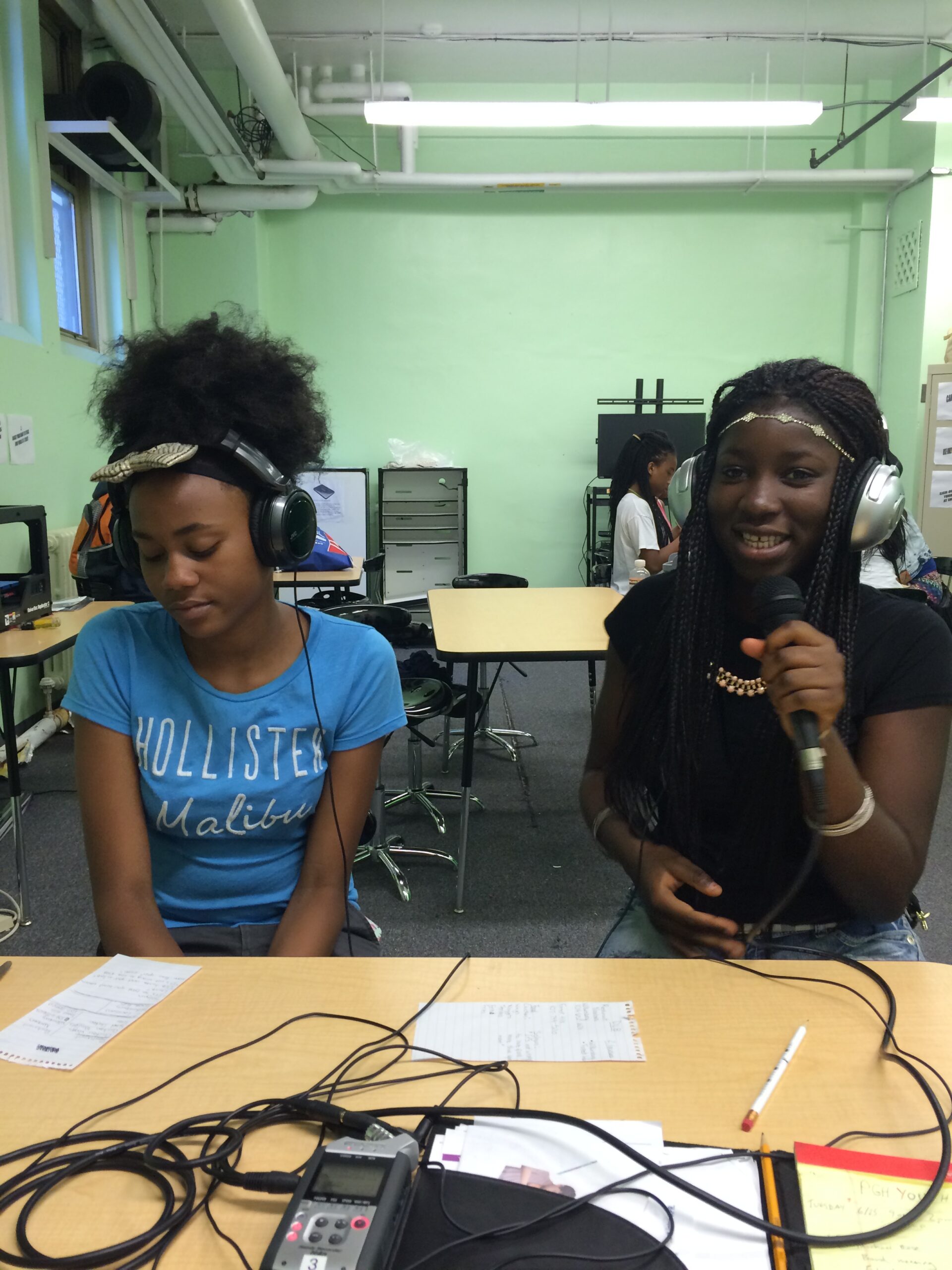 Two teens wearing headphones sitting at classroom desks, one holding and speaking into a microphone