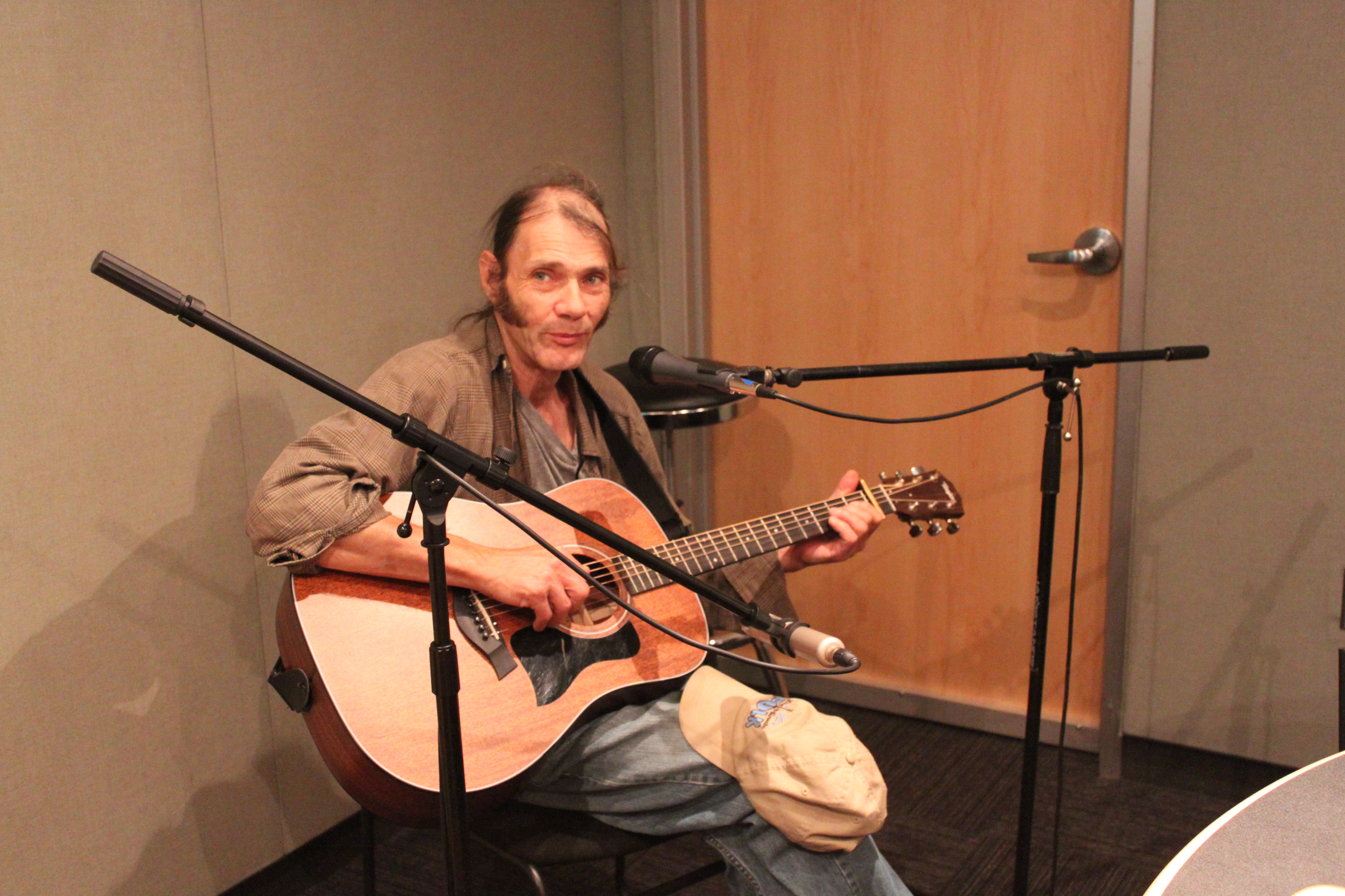 An adult playing an acoustic guitar sitting behind a microphone in a radio studio