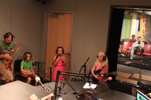 An adult holding a guitalele, a youth holding a guitalele, a youth standing behind a microphone, and a youth holding an electric guitar, all wearing headphones in a radio studio