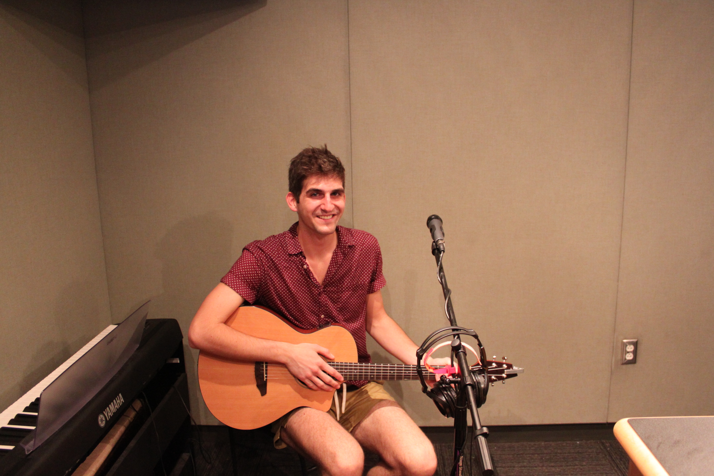 An adult holding an acoustic guitar sitting behind a microphone in a radio studio