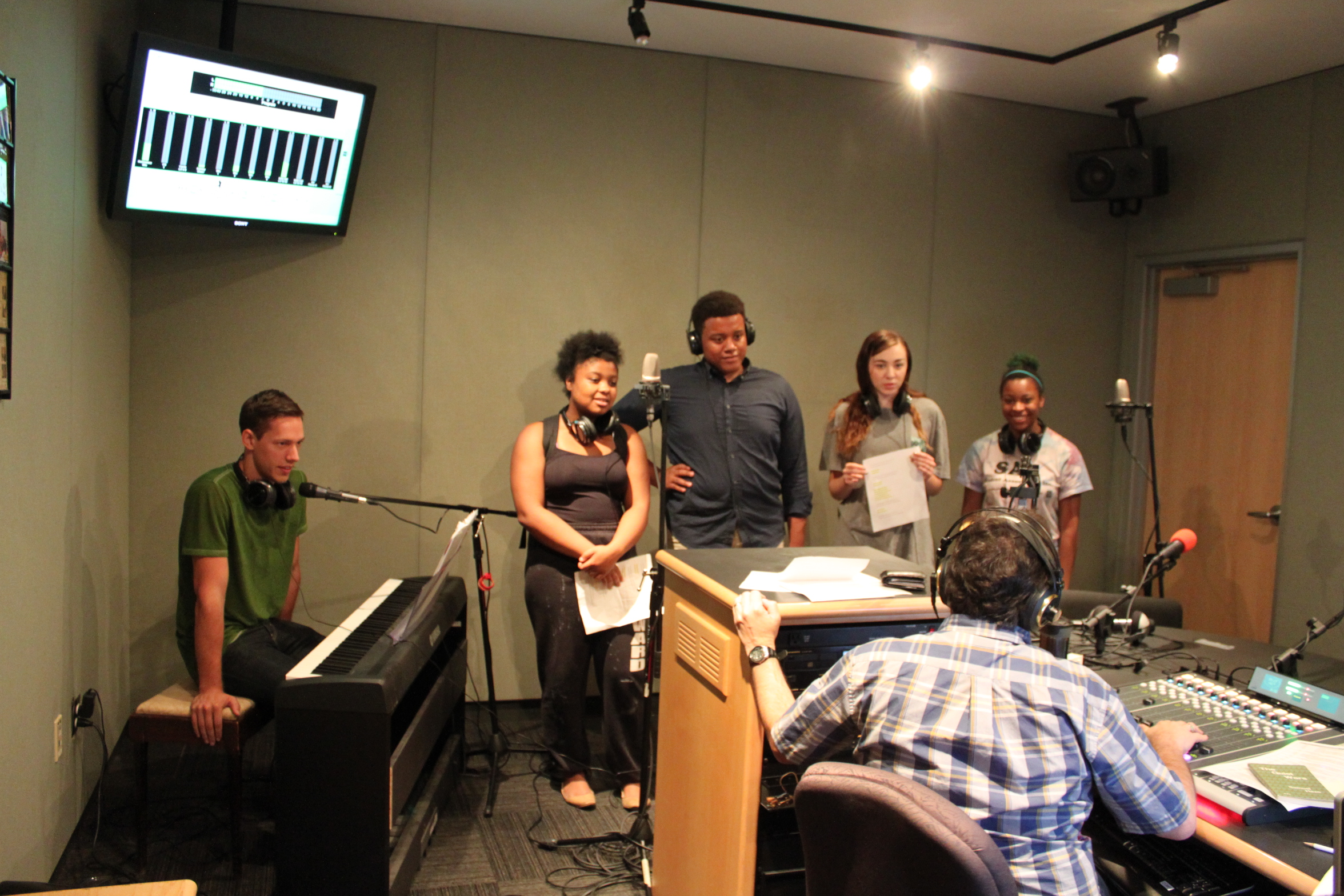 A teen sitting behind a piano keyboard and a microphone and four teens standing behind microphones being interviewed by an adult wearing headphones sitting behind a mixing board in a radio studio
