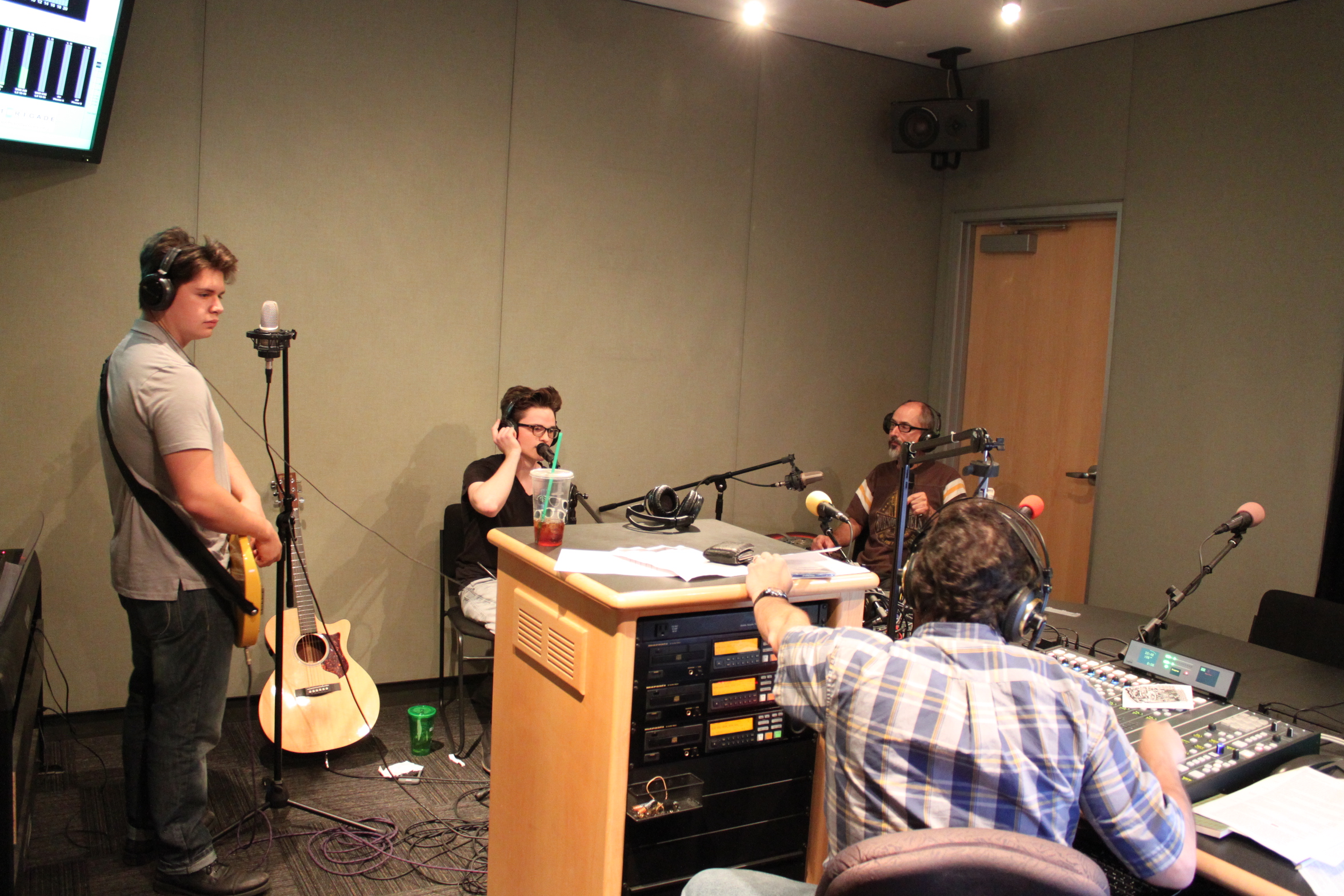 Two teens and an adult wearing headphones, one teen holding an electric string instrument, being interviewed by an adult wearing headphones sitting behind a mixing board in a radio studio