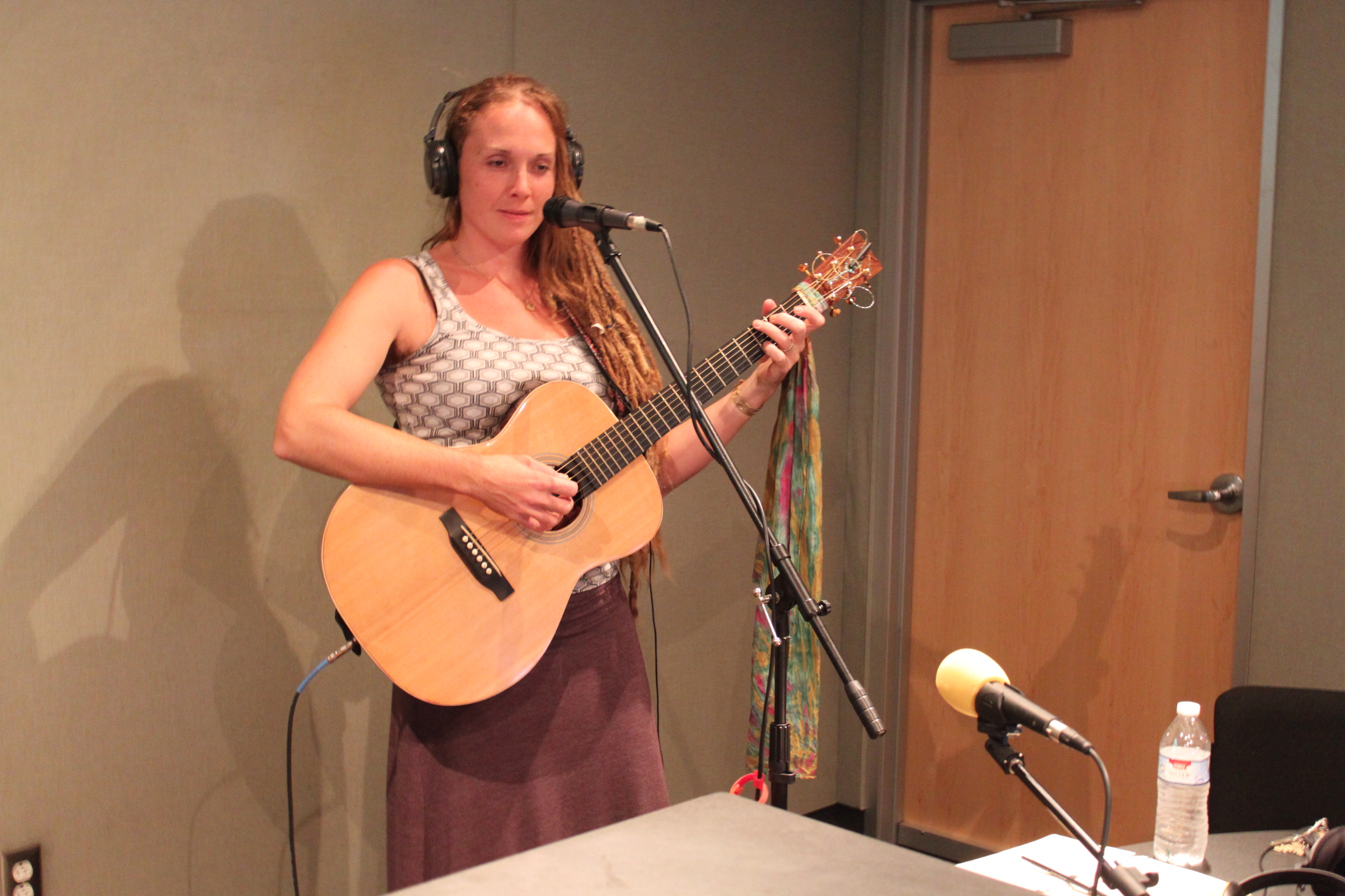 An adult wearing headphones playing an acoustic guitar standing behind a microphone in a radio studio