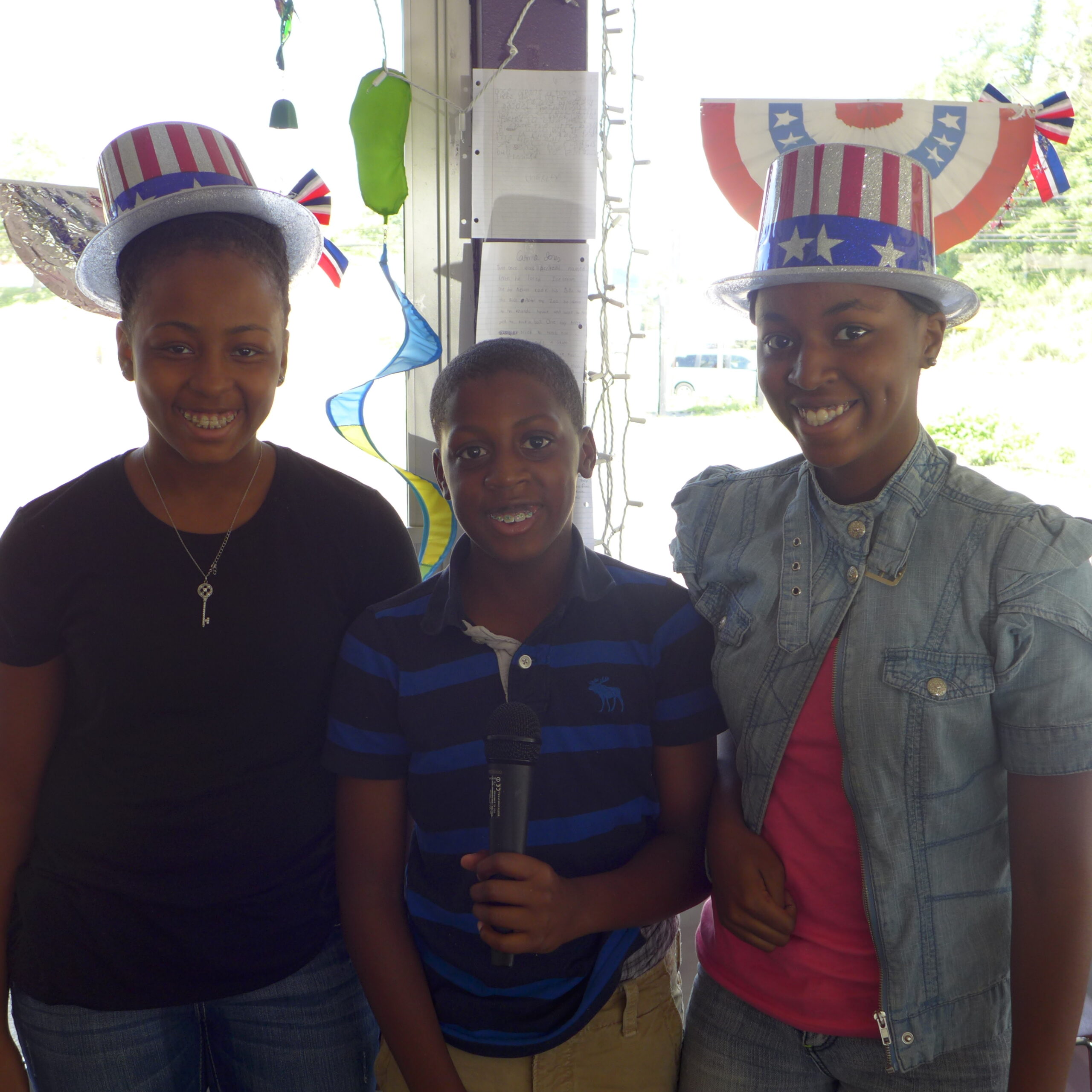 Two youth wearing american flag styled hats and a youth holding a microphone, all smiling in profile