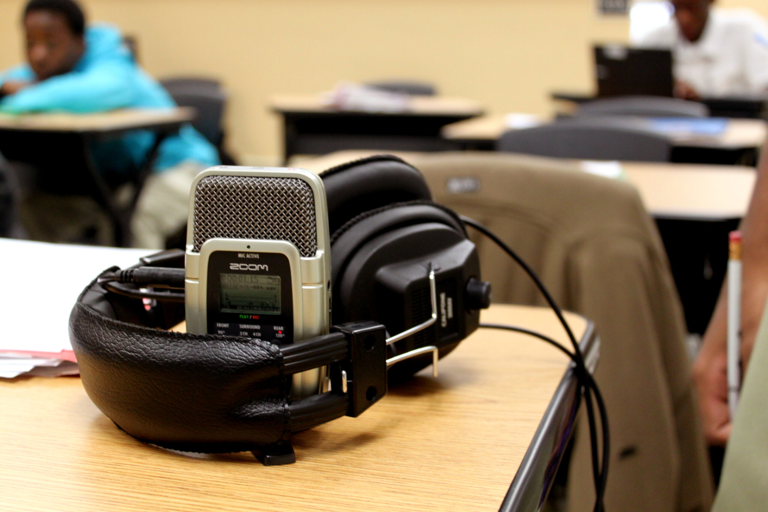 A pair of headphones around a portable recorder sitting on a wooden desk in a classroom