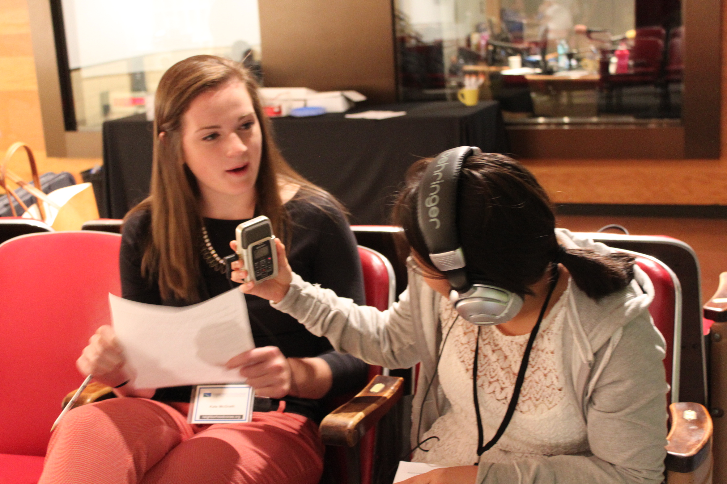 A teen wearing headphones holding a portable recorder up to another teen holding a piece of paper