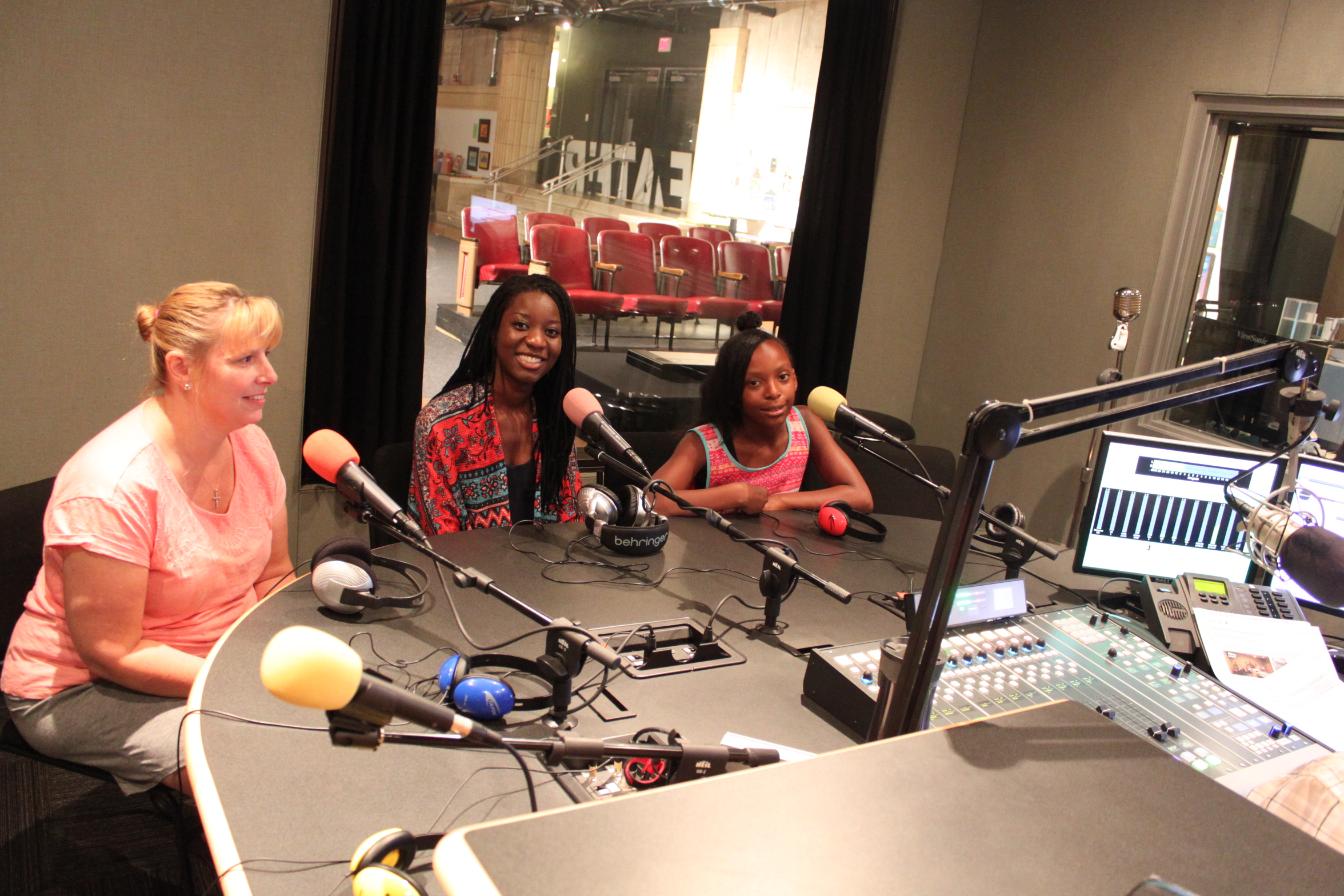 A youth, a teen, and an adult smiling and sitting behind microphones in a radio studio