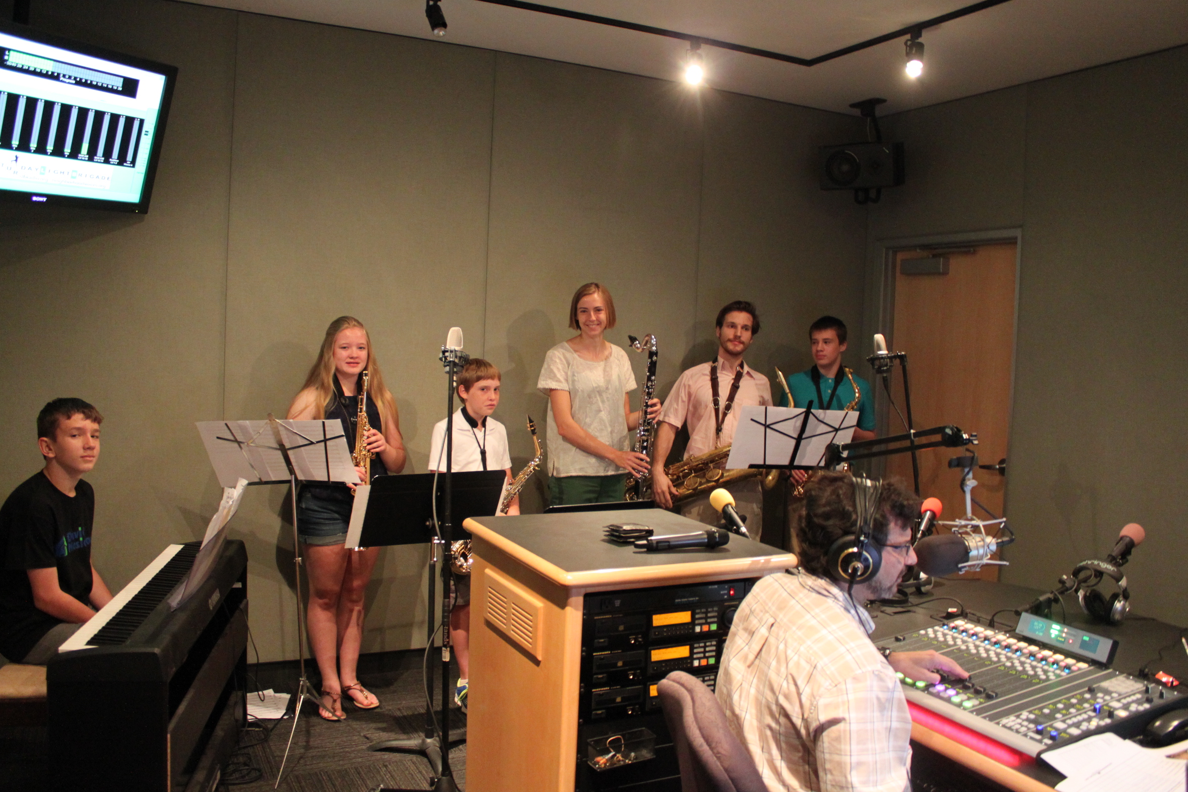 A youth sitting behind a piano keyboard, a youth holding a soprano saxophone, a youth holding an alto saxophone, a youth holding a bass clarinet, an adult holding a baritone saxophone, a youth holding an alto saxophone, and an adult wearing headphones sitting behind a microphone and a mixing board in a radio studio