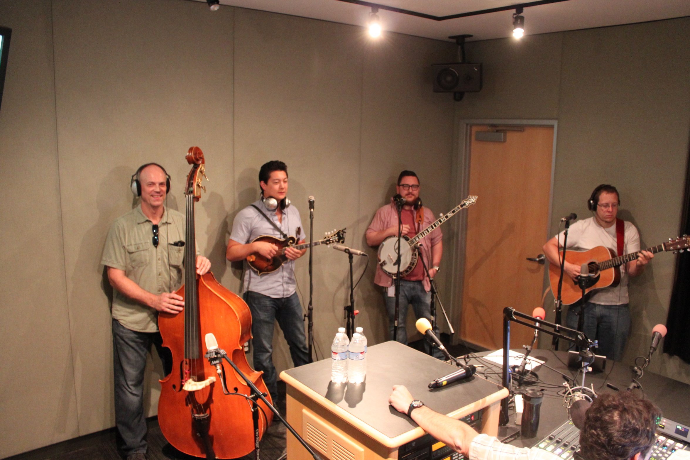 An adult wearing headphones holding an upright bass, an adult playing a mandolin, an adult holding a banjo, and an adult wearing headphones playing an acoustic guitar in a radio studio