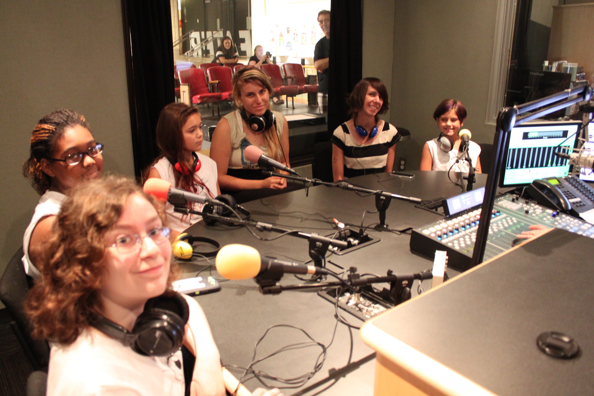 Six individuals wearing headphones sitting behind microphones in a radio studio