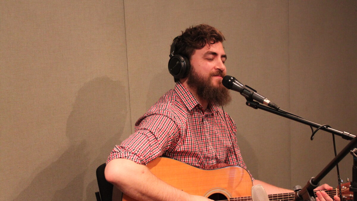 An adult wearing headphones playing acoustic guitar and singing into a microphone in a radio studio
