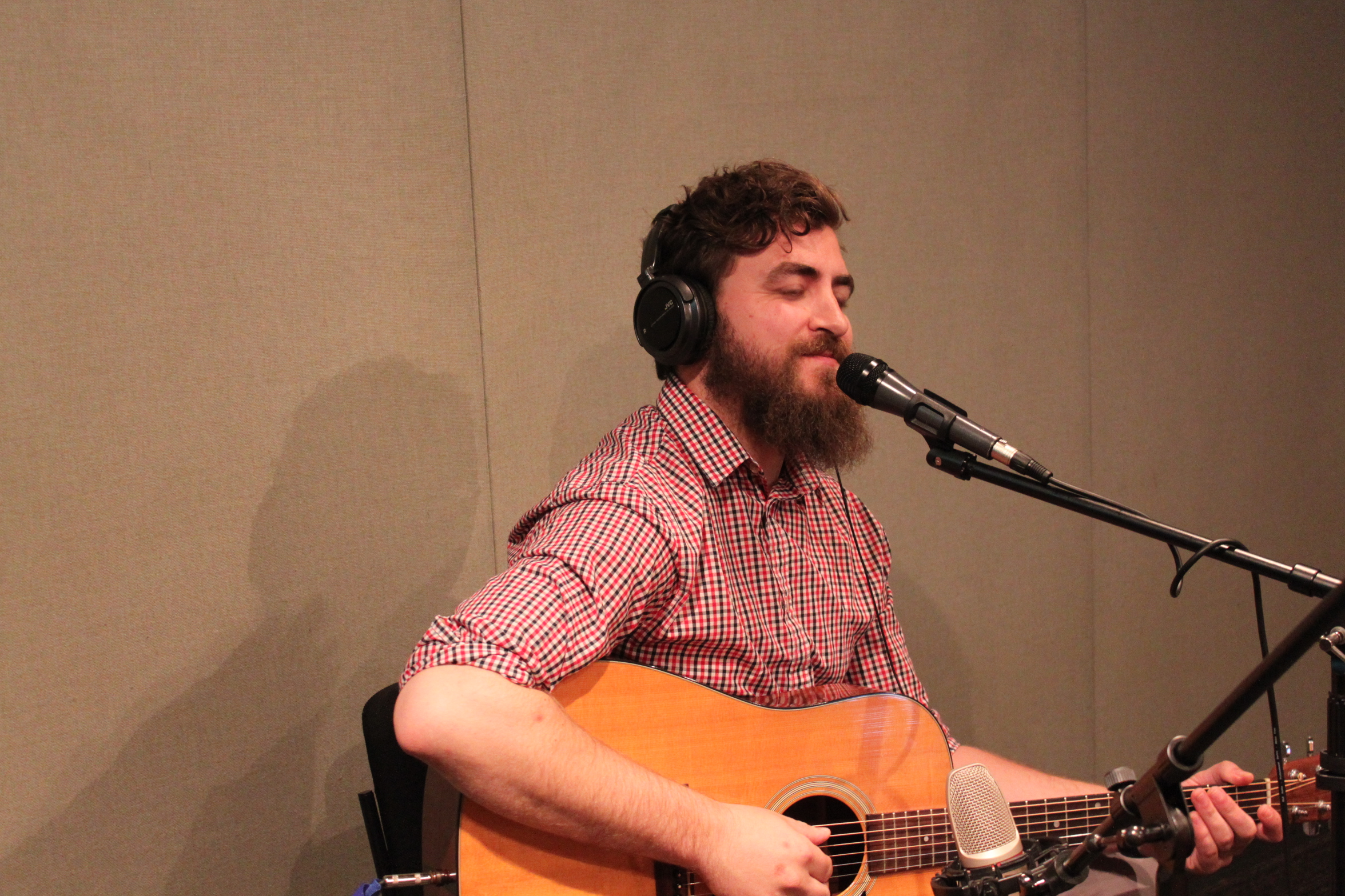 An adult wearing headphones playing acoustic guitar and singing into a microphone in a radio studio