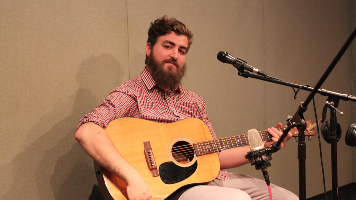 An adult holding an acoustic guitar sitting behind a microphone in a radio studio