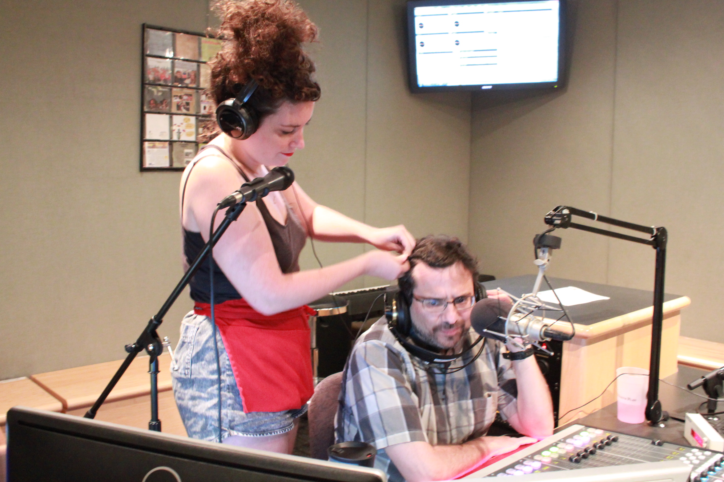 An adult wearing headphones standing behind a microphone braiding the hair of an adult sitting behind a microphone and a mixing board in a radio studio