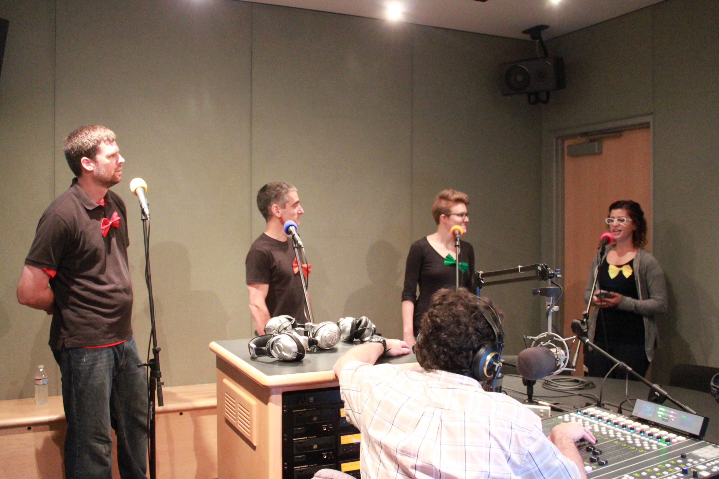 Four adults standing behind microphones and an adult wearing headphones sitting behind a microphone and a mixing board in a radio studio