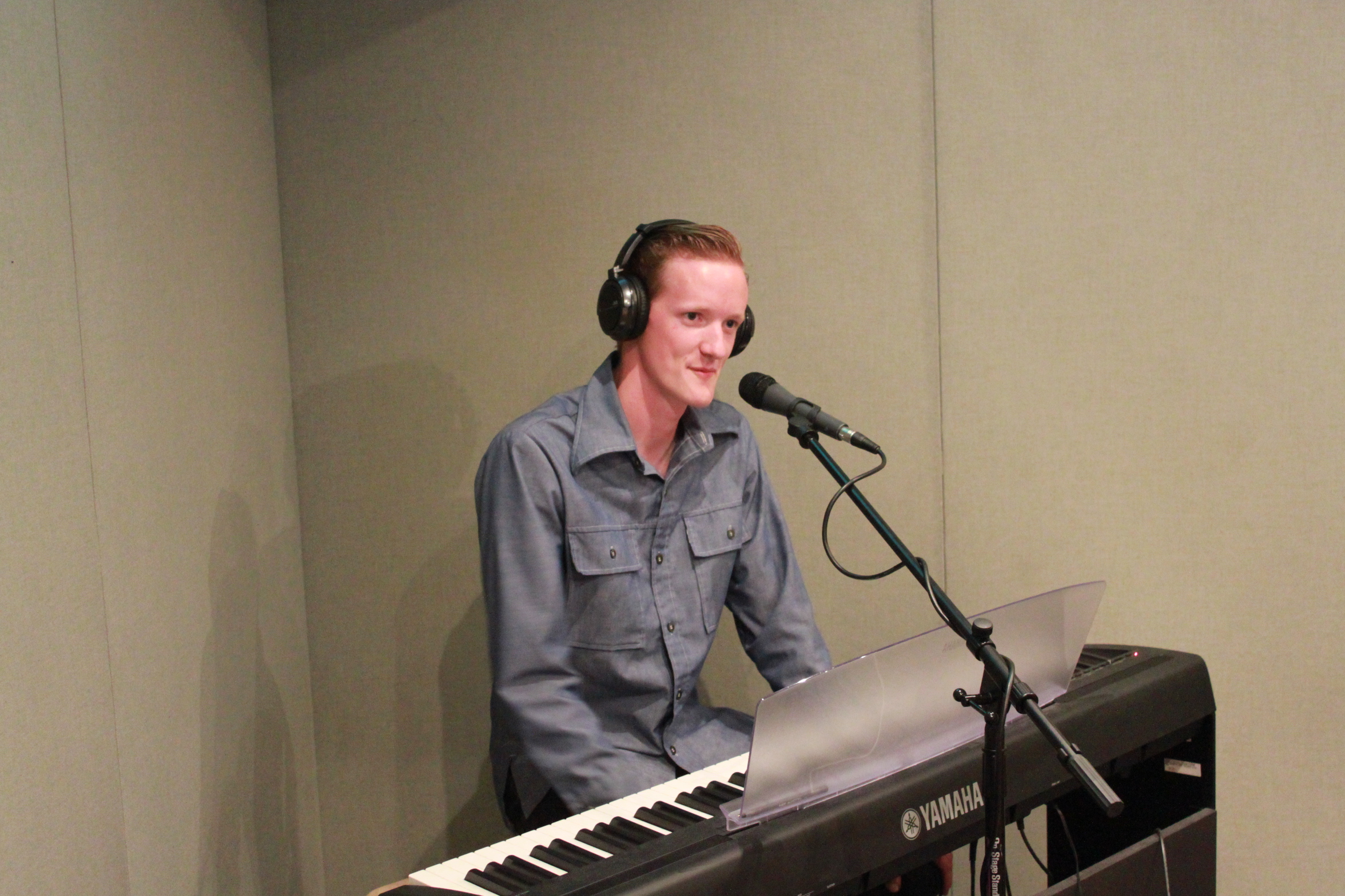 An adult wearing headphones sitting at a piano keyboard speaking into a microphone in a radio studio