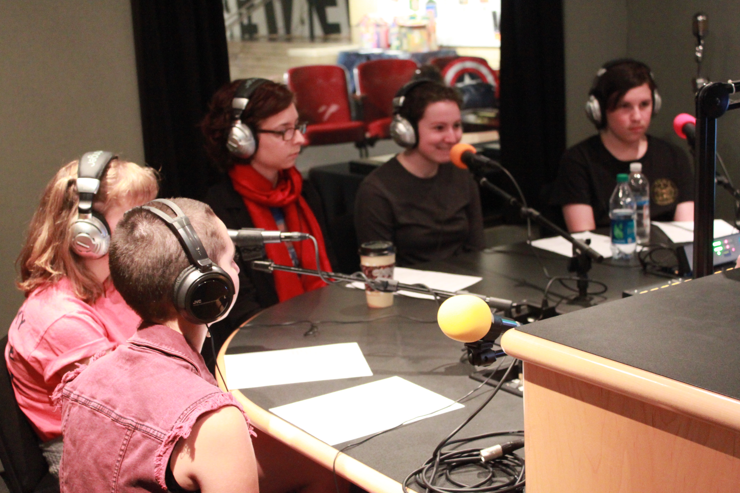Five teens wearing headphones speaking into microphones in a radio studio