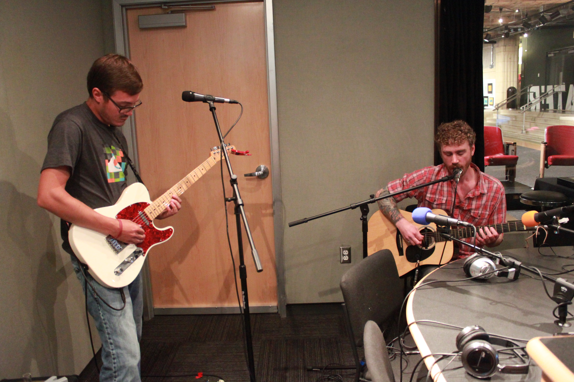 An adult playing electric guitar and an adult playing acoustic guitar and singing into a microphone in a radio studio