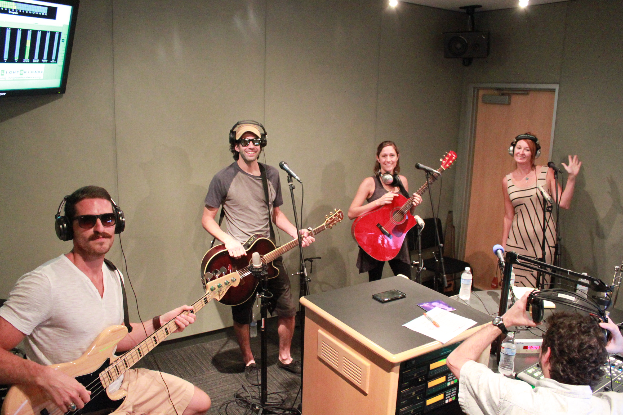 An adult wearing headphones holding an electric bass, an adult wearing headphones holding an acoustic guitar, an adult holding an acoustic guitar, and an adult wearing headphones waving at the camera in a radio studio