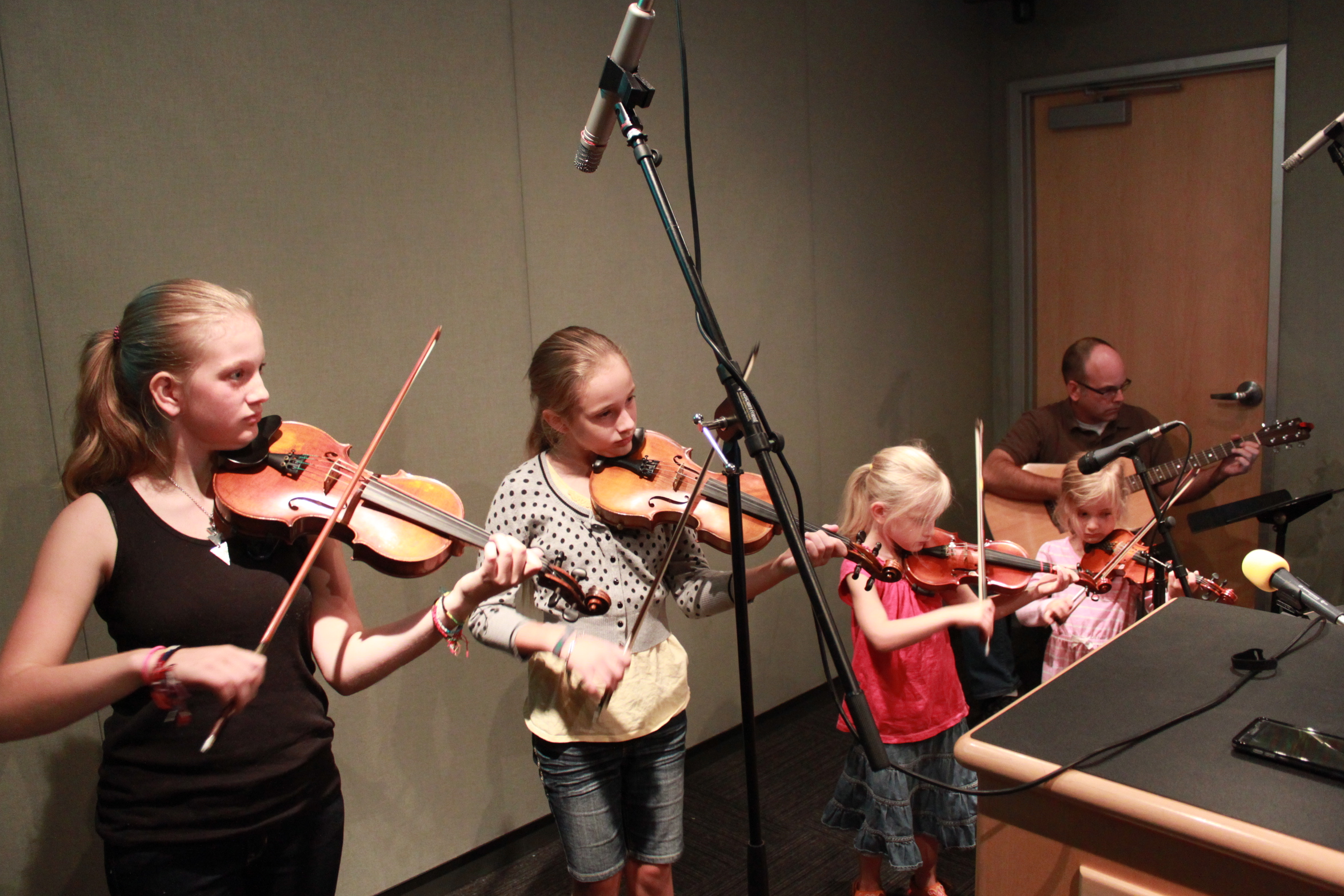 Four youth playing violins and an adult playing acoustic guitar in a radio studio