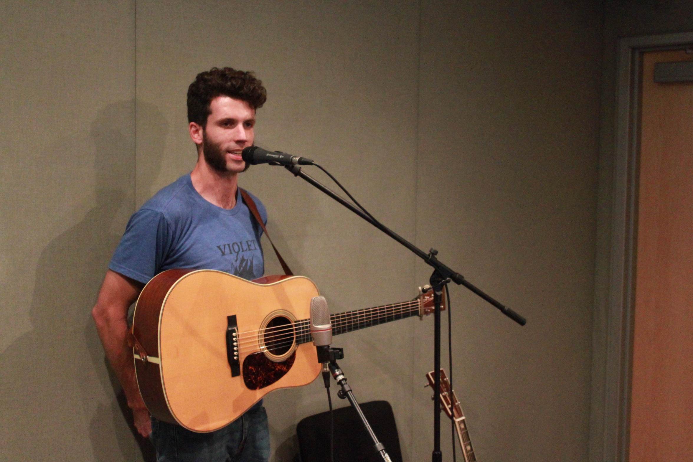 An adult holding an acoustic guitar speaking into a microphone in a radio studio