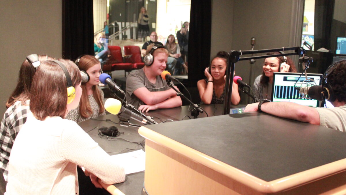 Six teens wearing headphones speaking into microphones being interviewed by an adult wearing headphones sitting behind a microphone in a radio studio
