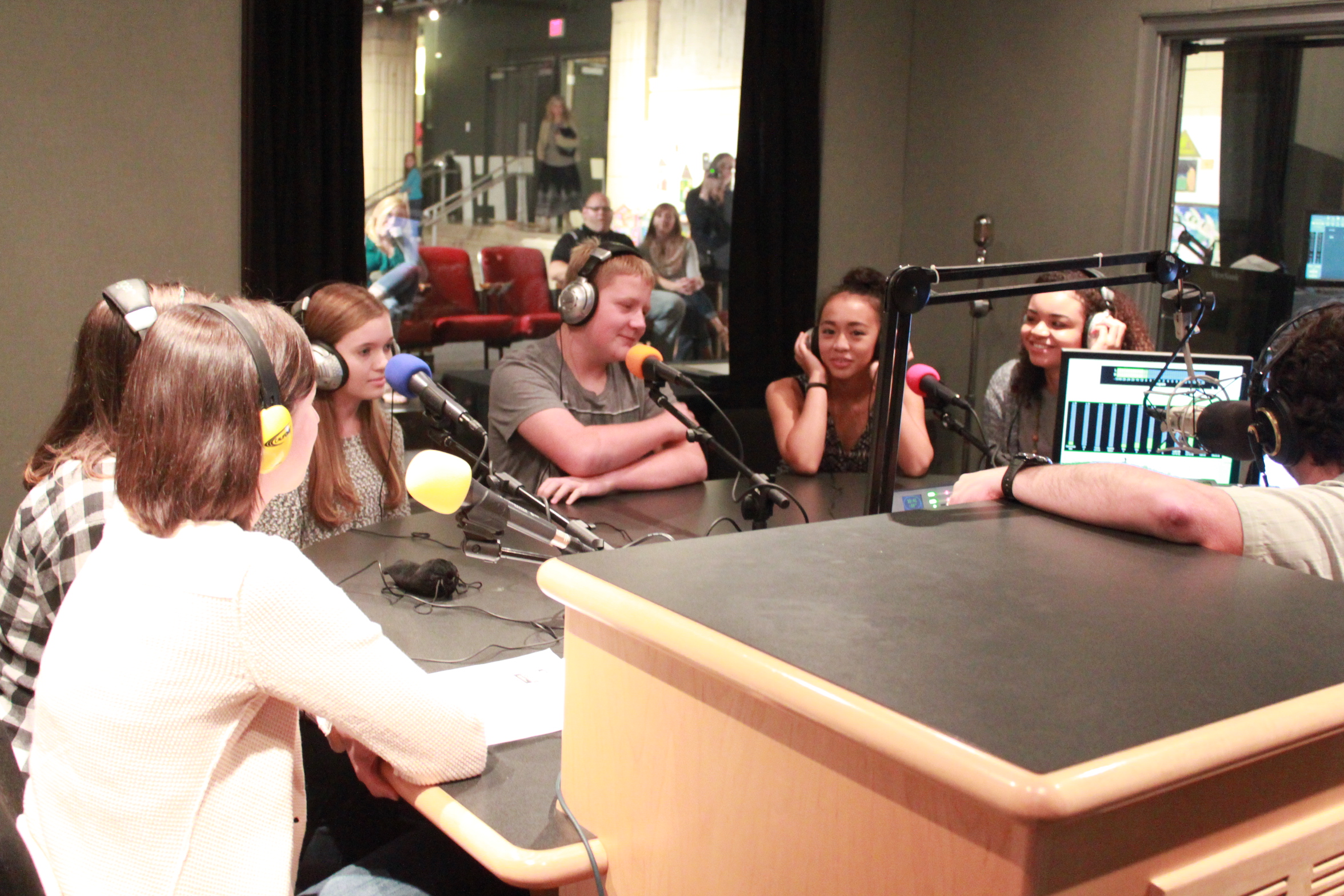 Six teens wearing headphones speaking into microphones being interviewed by an adult wearing headphones sitting behind a microphone in a radio studio