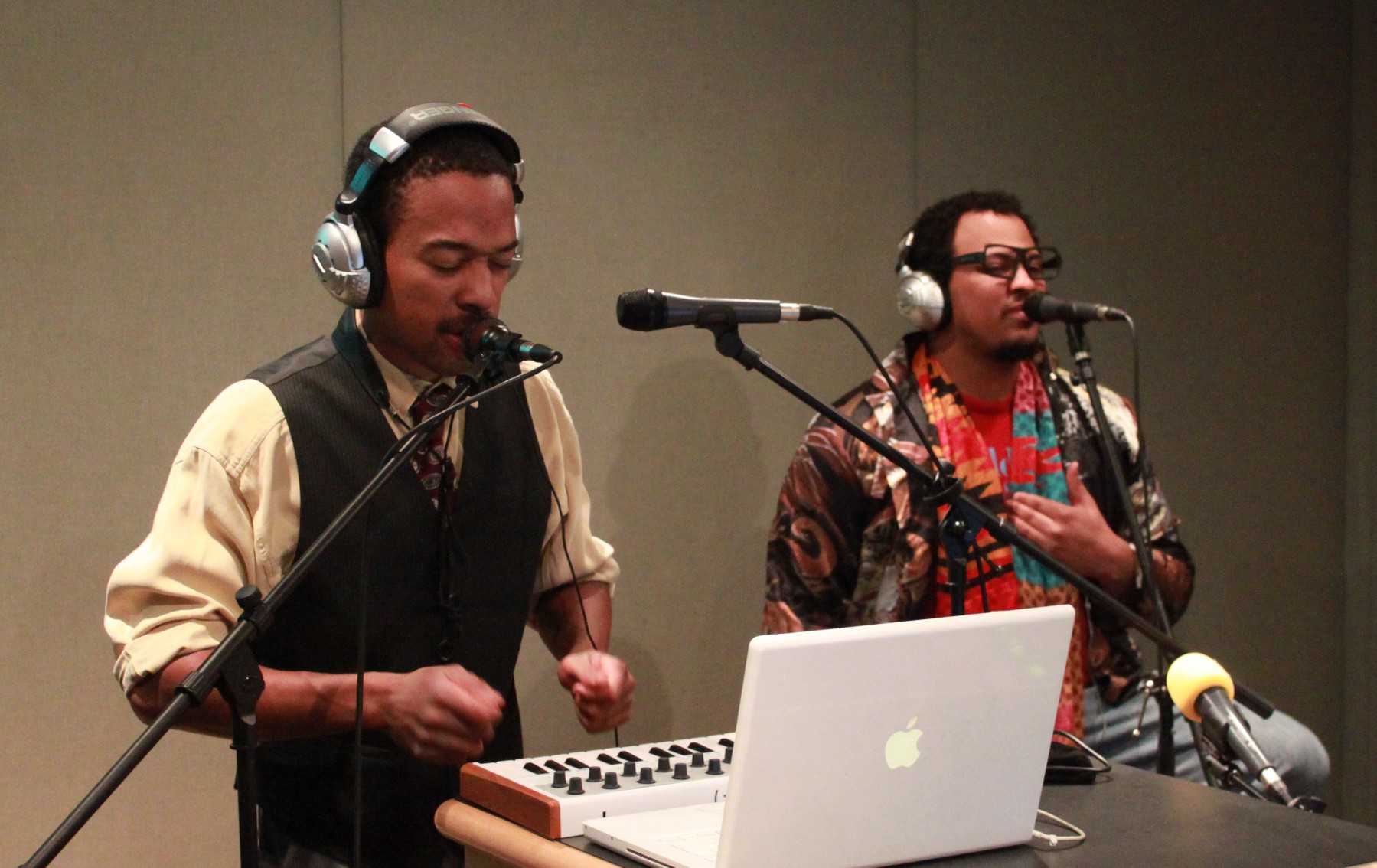 Two adults wearing headphones singing into microphones in a radio studio, one interacting with a small piano keyboard connected to a laptop