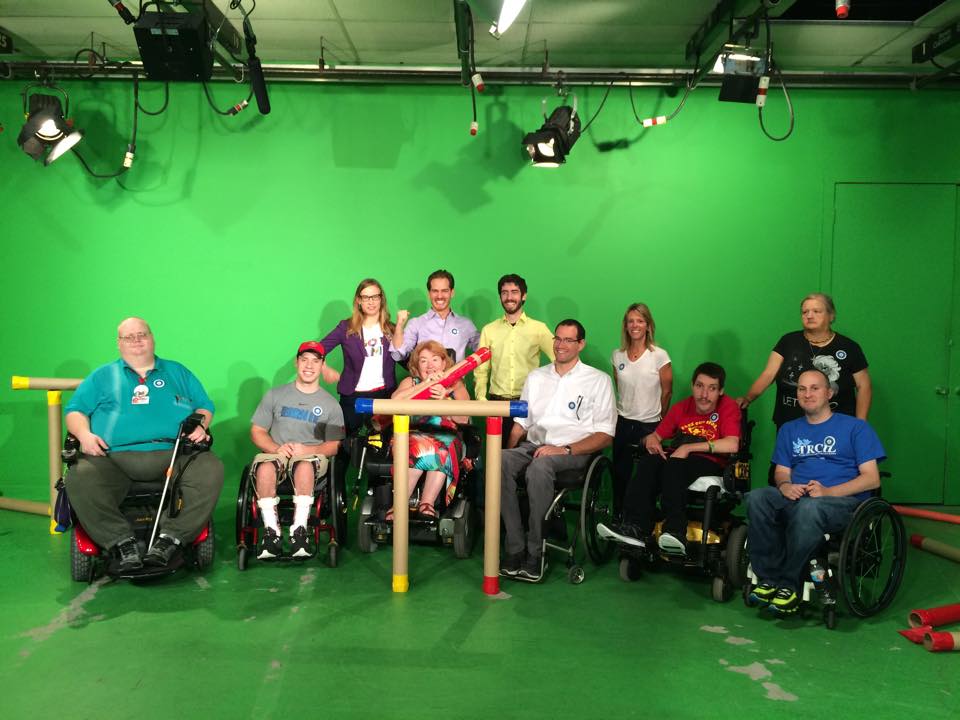 Eleven individuals, 6 sitting in wheelchairs, smiling and posing in a green room