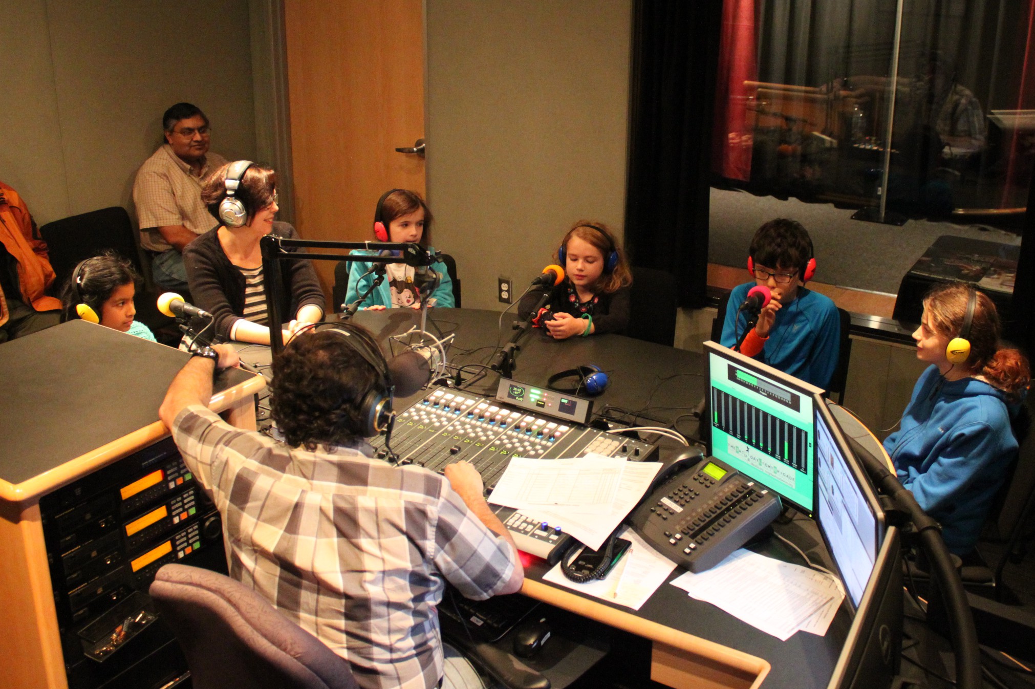 Five youth and an adult wearing headphones speaking into microphones being interviewed by an adult wearing headphones sitting behind a microphone and a mixing board as another adult listens in the background in a radio studio
