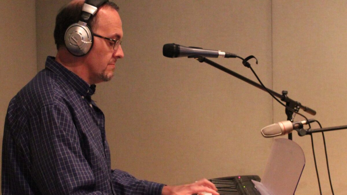 IMG_0189 An adult wearing headphones playing a piano keyboard and sitting behind a microphone in a radio studio