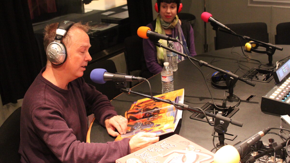 IMG_0190 Two adults wearing headphones in a radio studio, one looking down at a picture book and speaking into a microphone