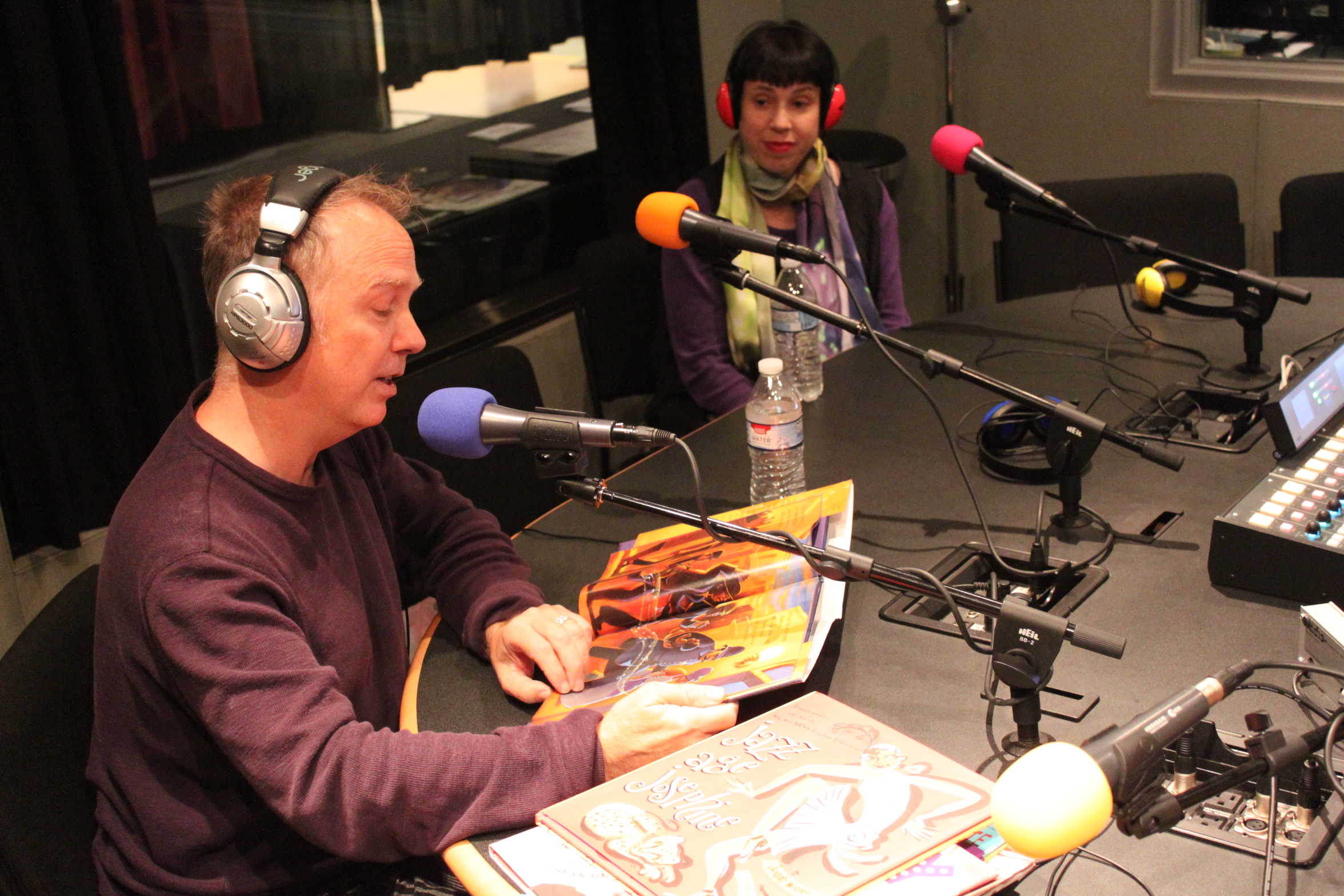 Two adults wearing headphones in a radio studio, one looking down at a picture book and speaking into a microphone