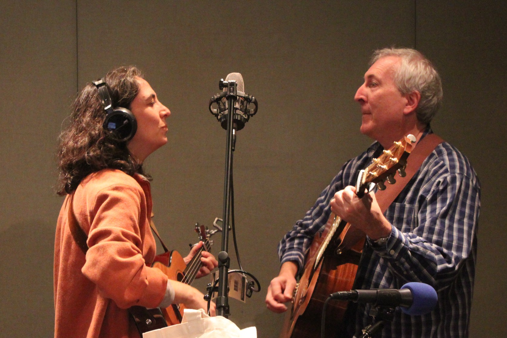 An adult playing a ukulele and an adult playing an acoustic guitar, both singing into a microphone in a radio studio
