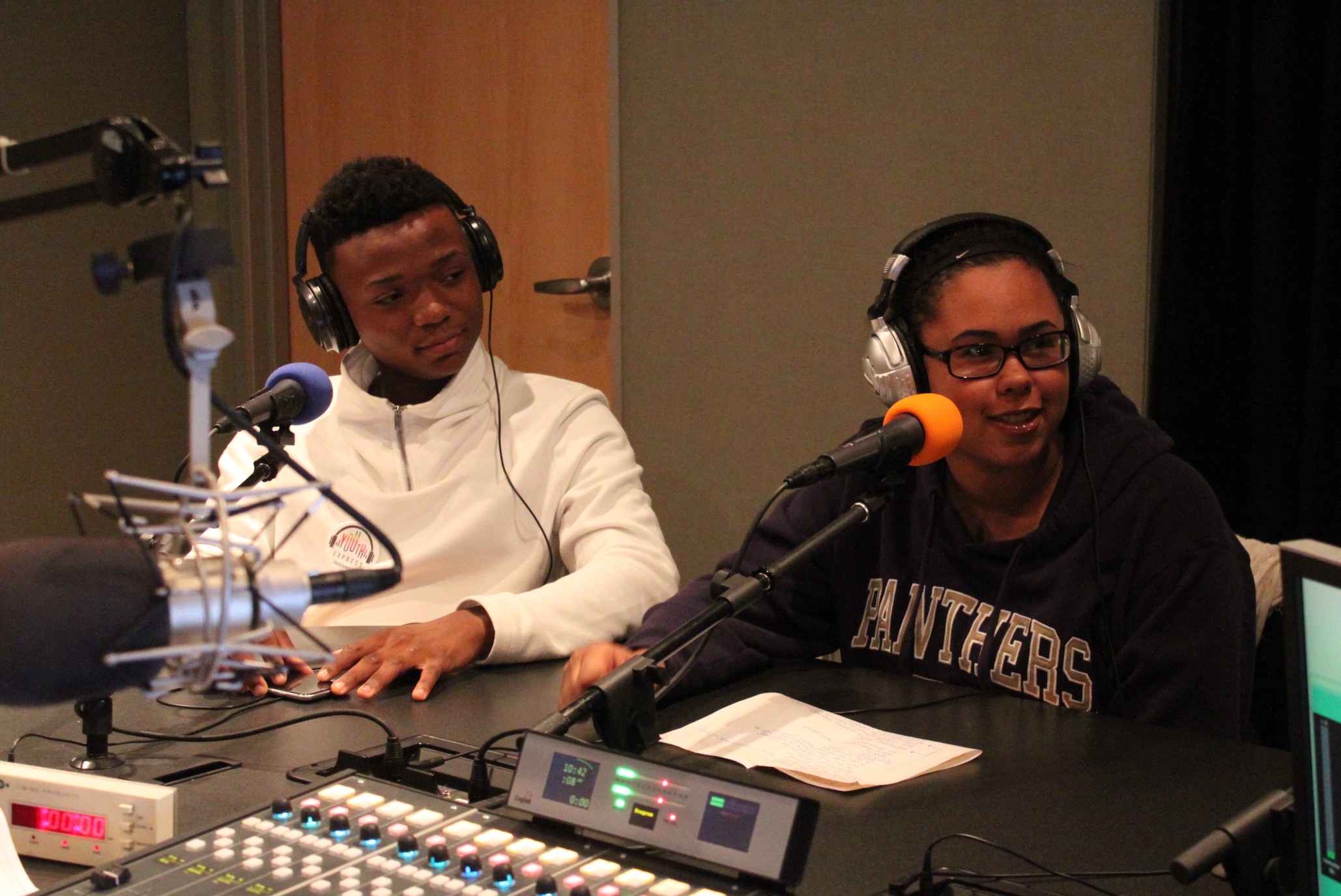 Two teens wearing headphones speaking into microphones in a radio studio