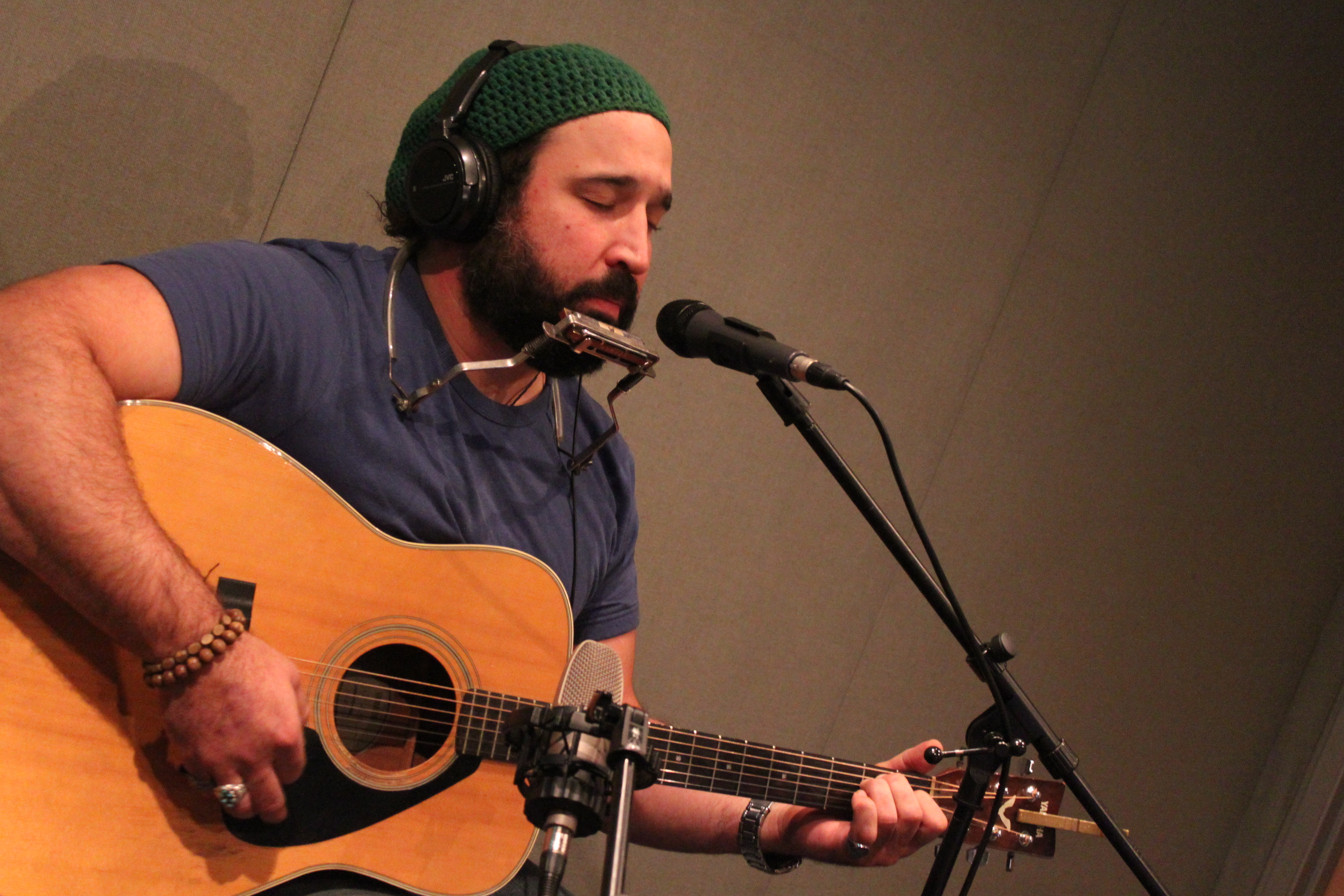 An adult wearing headphones playing acoustic guitar and singing into a microphone in a radio studio