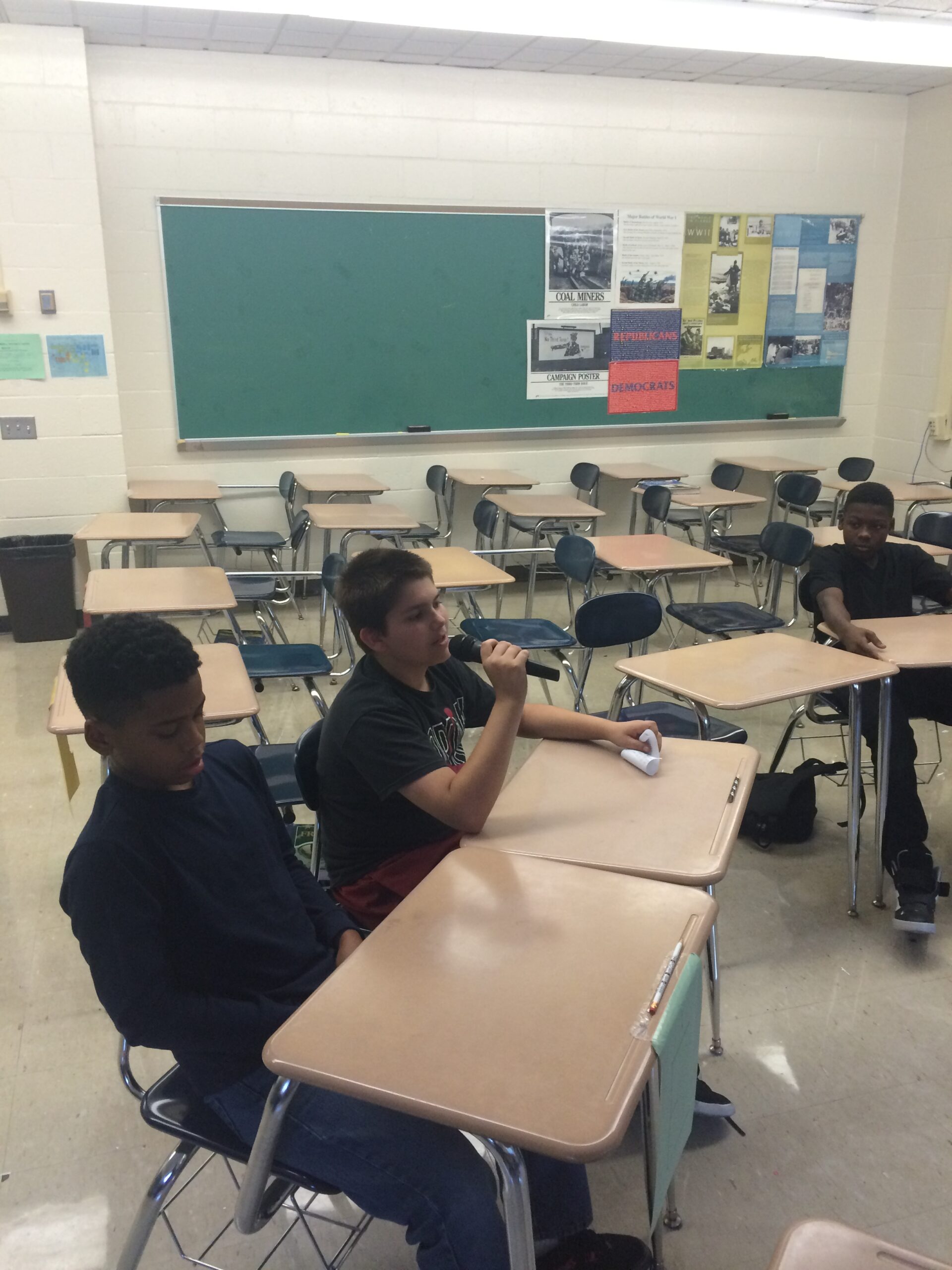 Three youth sitting at classroom desks, one holding and speaking into a microphone