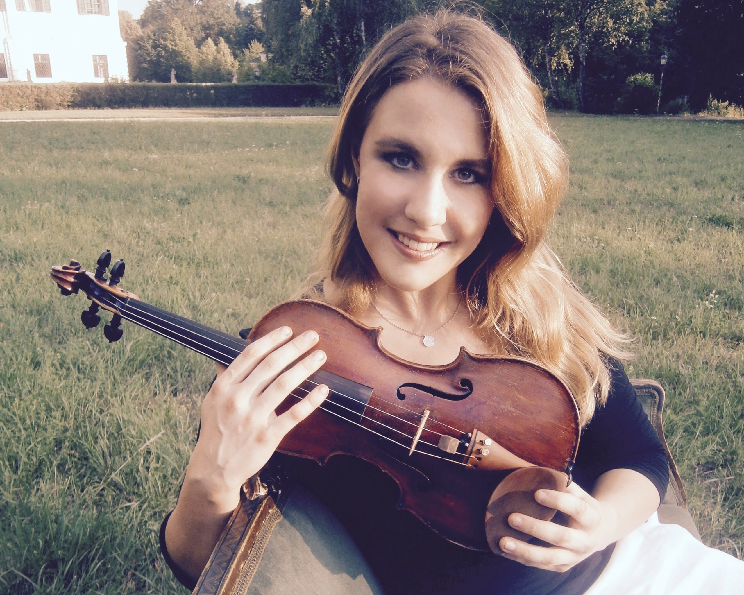 A teen holding a violin standing in a large green field of grass