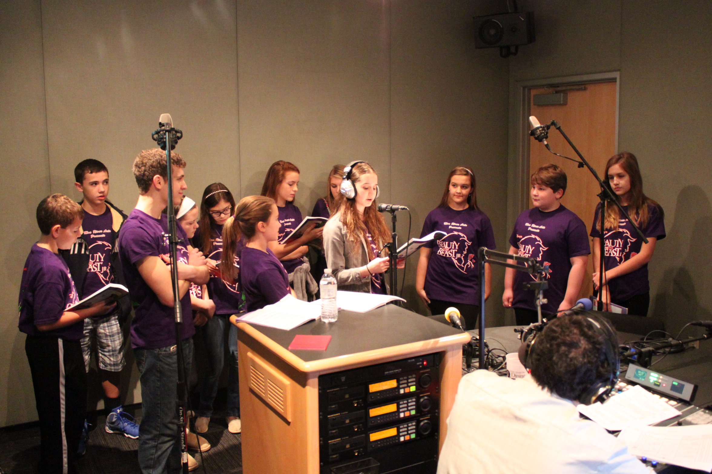 Twelve youth standing behind microphones reading from scripts as an adult wearing headphones sitting behind a microphone and a mixing board listens in a radio studio