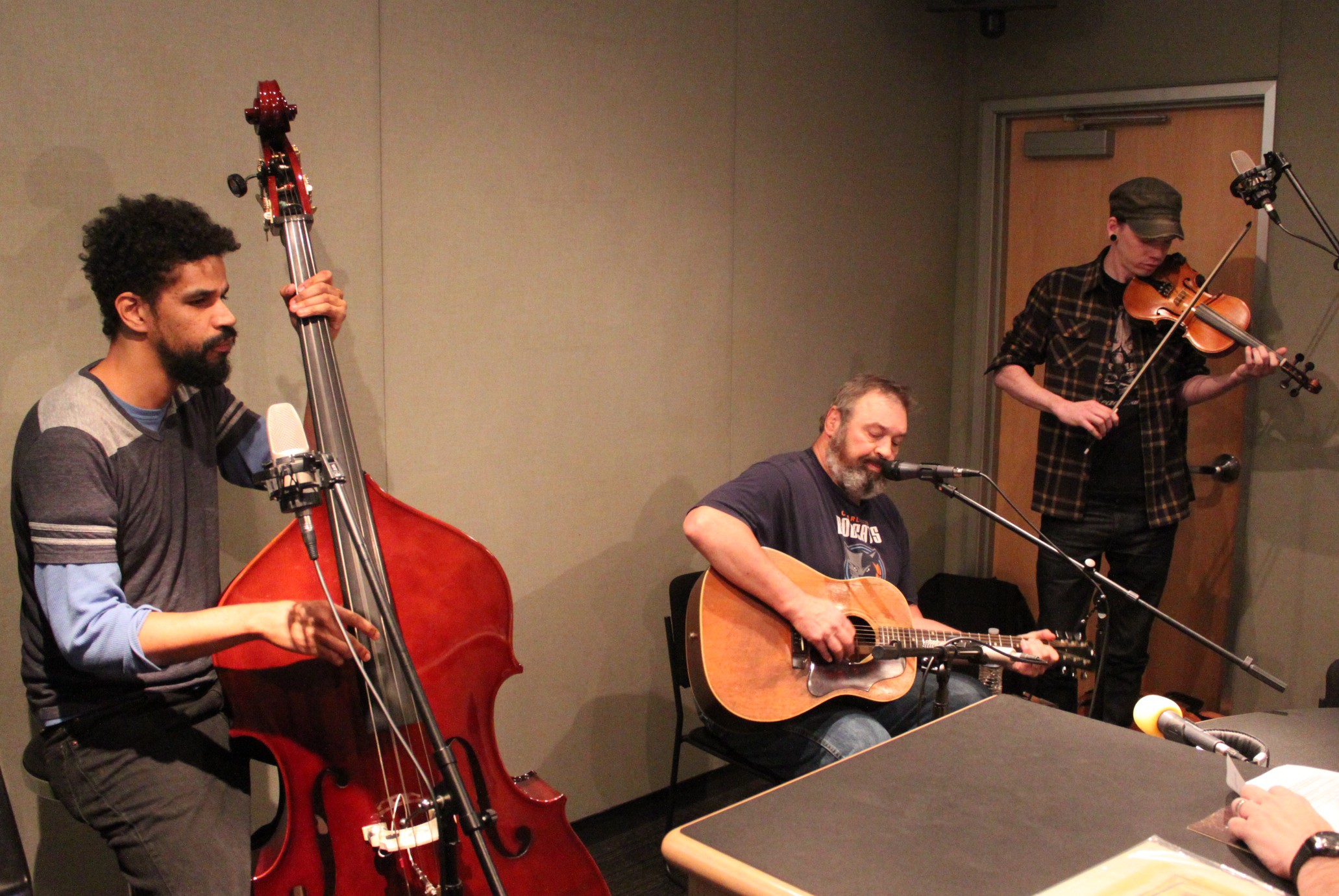 An adult playing upright bass, an adult playing acoustic guitar and singing into a microphone, and an adult playing fiddle in a radio studio