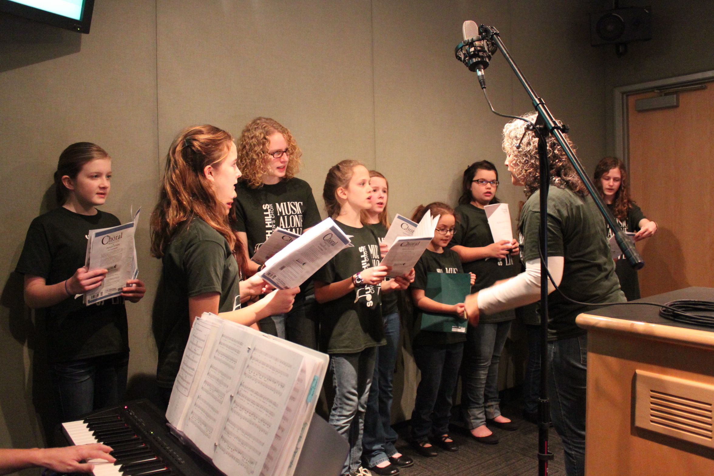 Ten youth singing in a choir being conducted by an adult in a radio studio