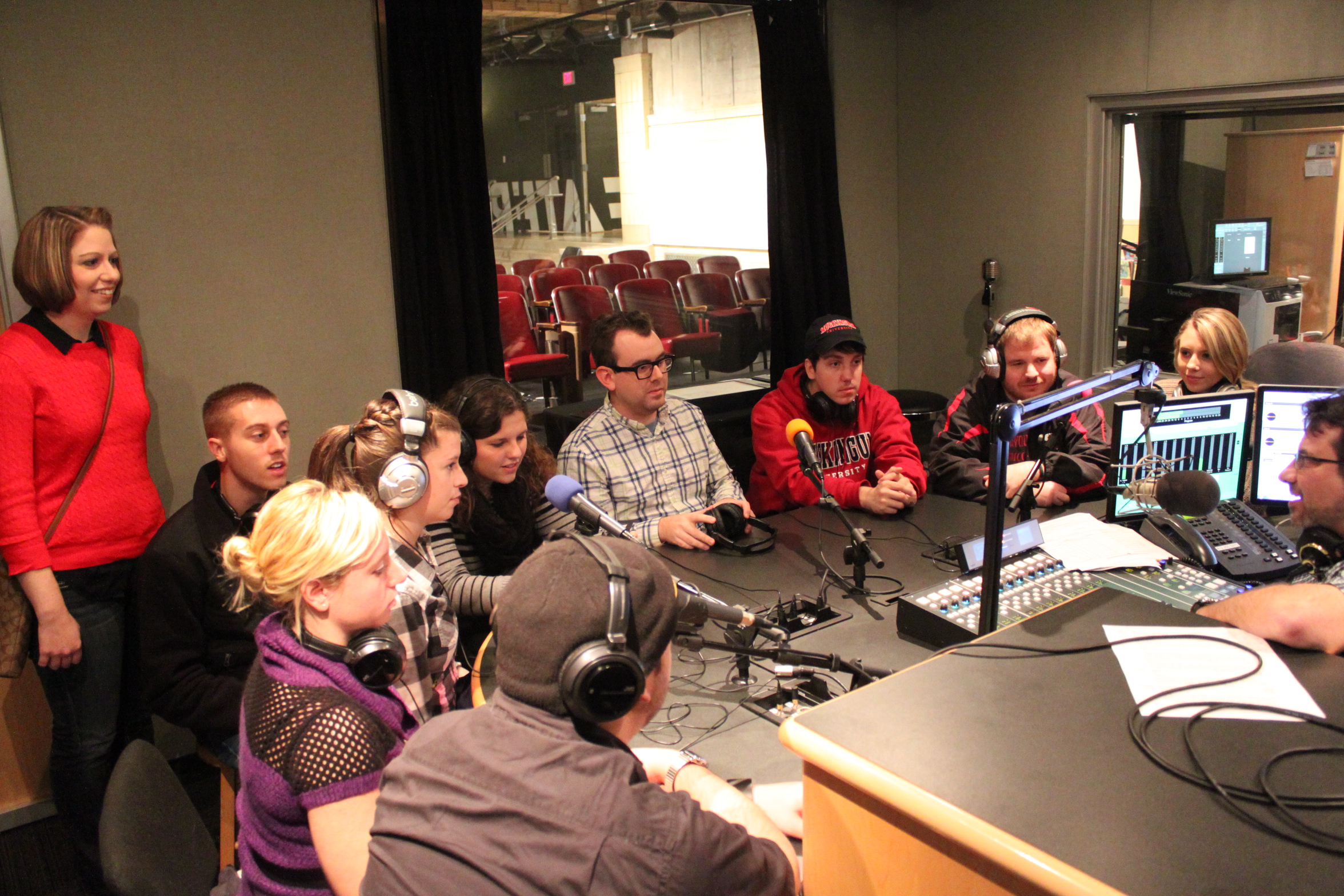 Ten adults, five wearing headphones, sitting behind microphones being interviewed by an adult sitting behind a microphone and a mixing board in a radio studio