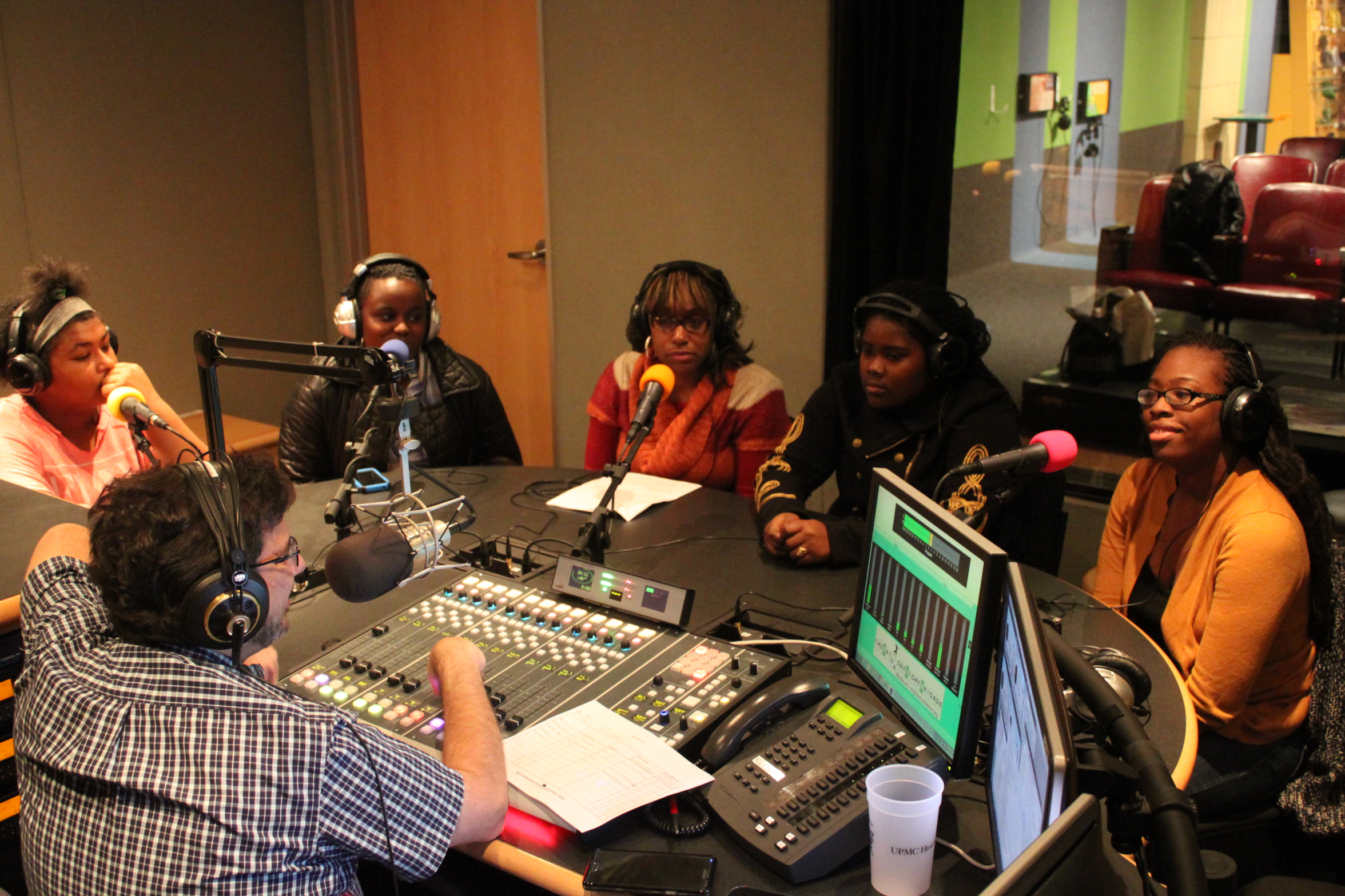 Five teens wearing headphones speaking into microphones being interviewed by an adult wearing headphones sitting behind a microphone and a mixing board in a radio studio