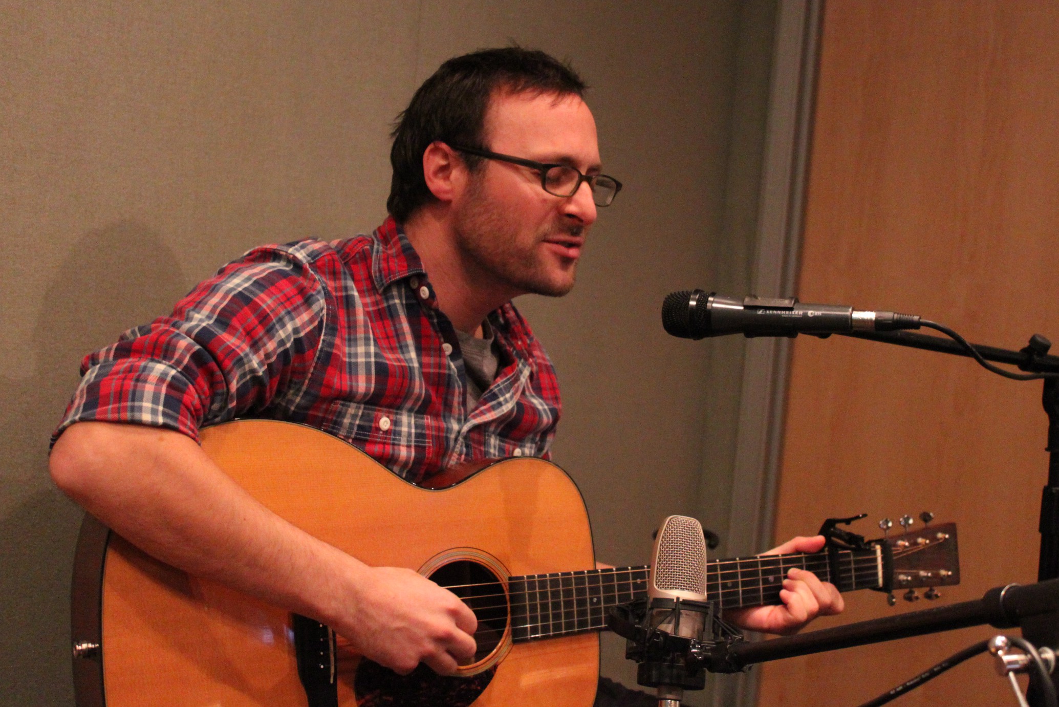 An adult playing acoustic guitar and singing into a microphone in a radio studio