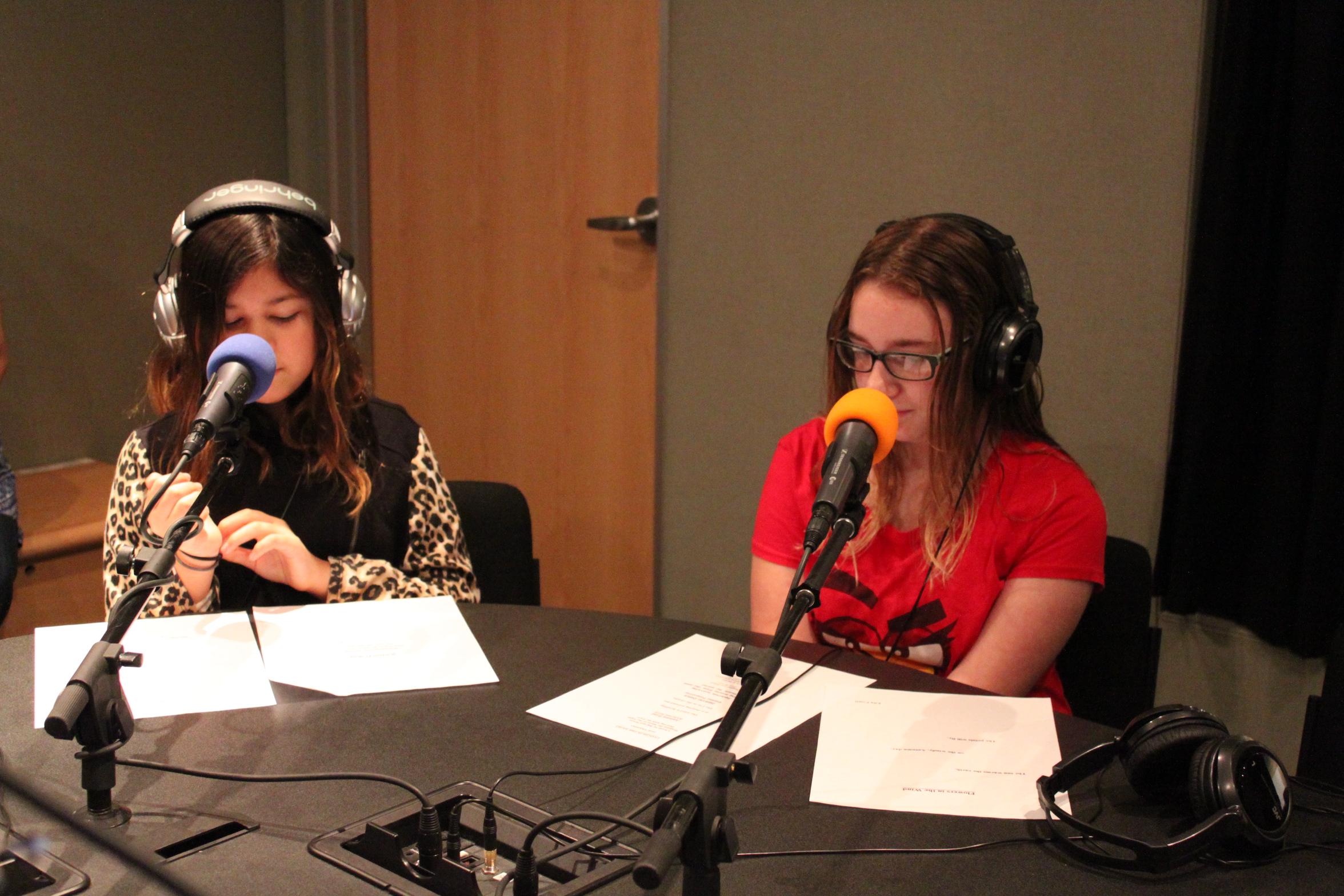 Two teens wearing headphones speaking into microphones and looking down at pieces of paper in a radio studio