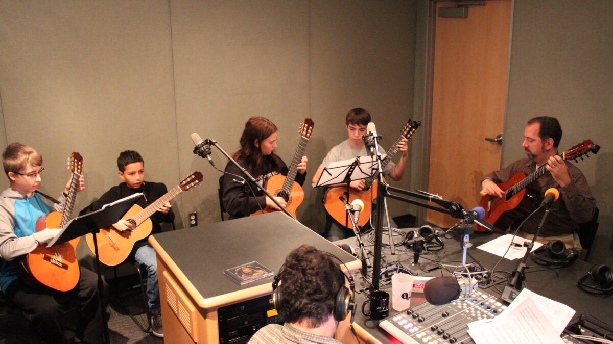 Four youth and an adult playing acoustic guitars as another adult listening wearing headphones sitting behind a microphone and a mixing board in a radio studio