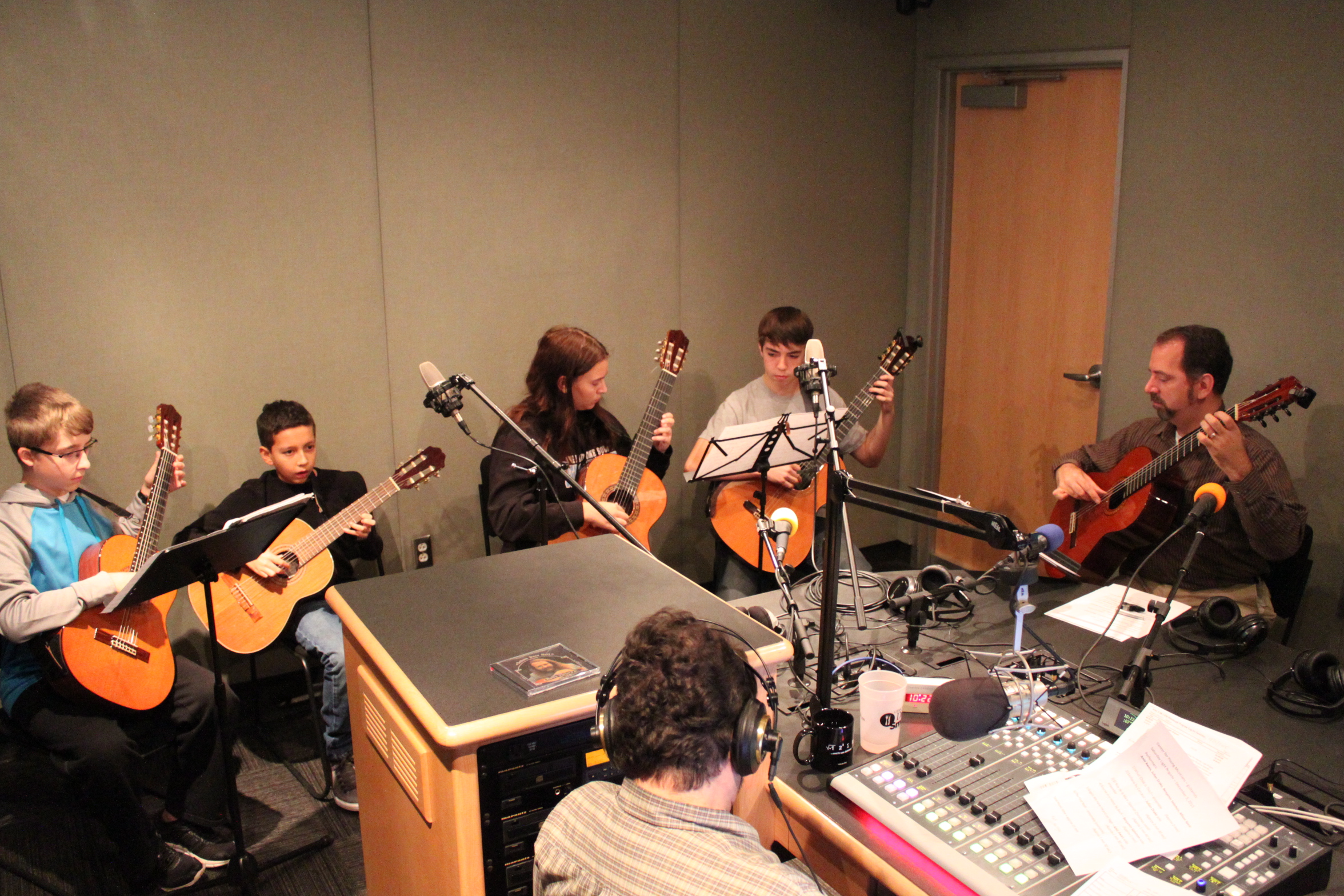 Four youth and an adult playing acoustic guitars as another adult listening wearing headphones sitting behind a microphone and a mixing board in a radio studio